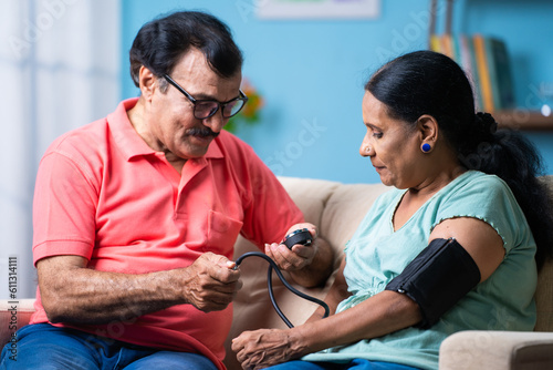 Fotografia Indian senior man checking blood pressure or bp of wife by using sphygmomanometer while sitting on sofa at home - concept of home health checkup, relationship and caring husband