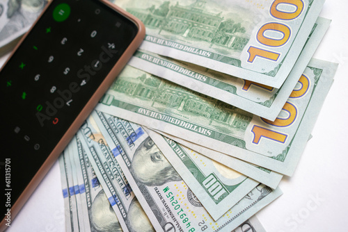 A stack of one hundred dollar bills and a calculator on a smartphone. A stack of one hundred dollar bills and a smartphone on a light background.