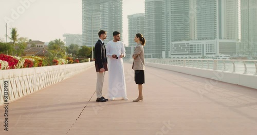 A man in traditional white Arab clothes talks with his European colleagues on the embankment of a modern city photo