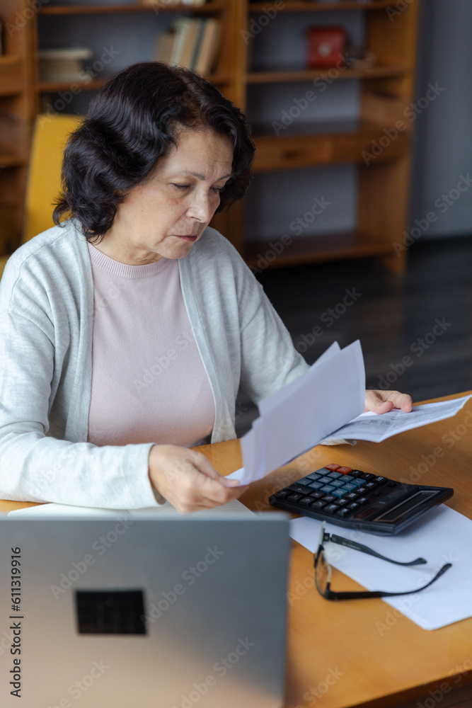 Middle aged senior woman sit with laptop and paper document. Pensive older mature lady reading paper bill pay online at home managing bank finances calculating taxes planning loan debt pension payment Middle aged senior woman sit with laptop and paper document. Pensive older mature lady reading paper bill pay online at home managing bank finances calculating taxes planning loan debt pension payment
