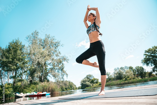 Indian woman practicing yoga and pilates standing one leg on wooden pier by river outdoors.