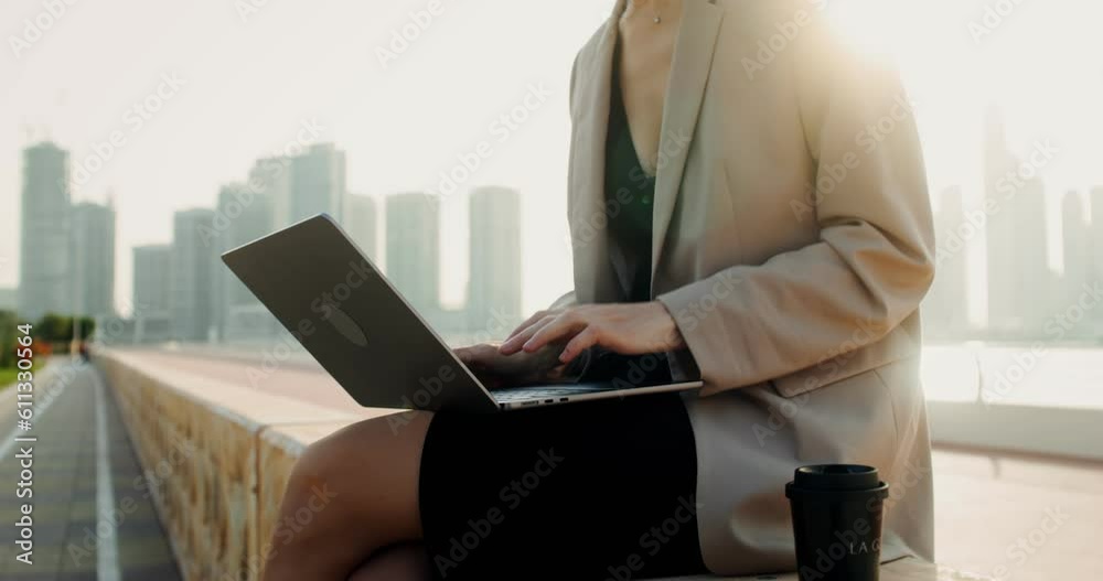A woman in business clothes uses a laptop while sitting on the embankment parapet. Close-up of female hands, no face