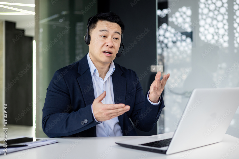 Portrait of young Asian male consultant in support service. Sitting in the office at the table, wearing headphones and a microphone, talking with clients online from a laptop Portrait of young Asian male consultant in support service. Sitting in the office at the table, wearing headphones and a microphone, talking with clients online from a laptop