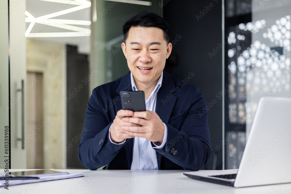 Young handsome Asian man, office worker sitting in the office at the table, fiddling with the phone, typing messages, chatting