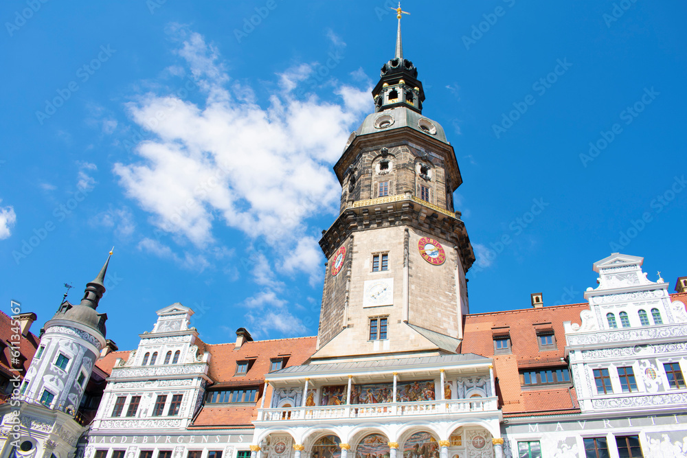 View of ancient building with large tower and spire, white walls and grey graphic paintings, windows and red roof. Old architecture, palace. Sunny day with blue sky. Dresden, Germany, May 2023.