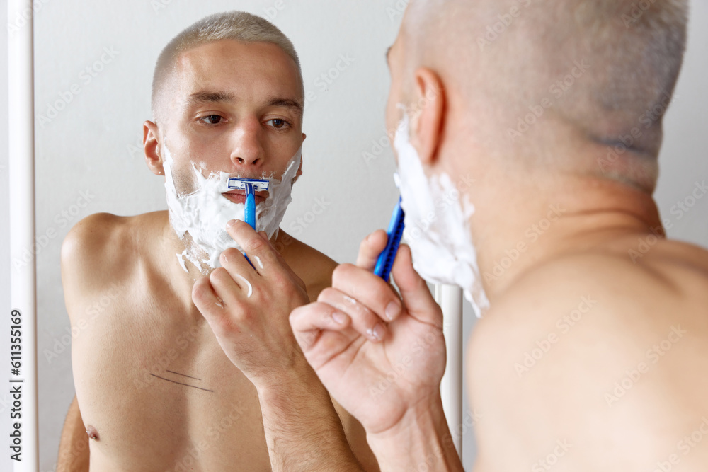 Portrait of young shirtless man shaving his beard, looking in mirror against white studio background. Concept of men's beauty, skincare, hygiene, cosmetology, health. Copy space for ad
