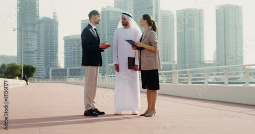 A man and a woman in European business clothes, and an Arab man in a traditional dress are talking standing outdoors. An Arab and a man shake hands photo