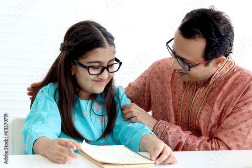 Cute Indian school student girl in traditional dress and glasses do homework with father  father teaching daughter kid at home  parent become teacher tutor involvement in childhood education in family
