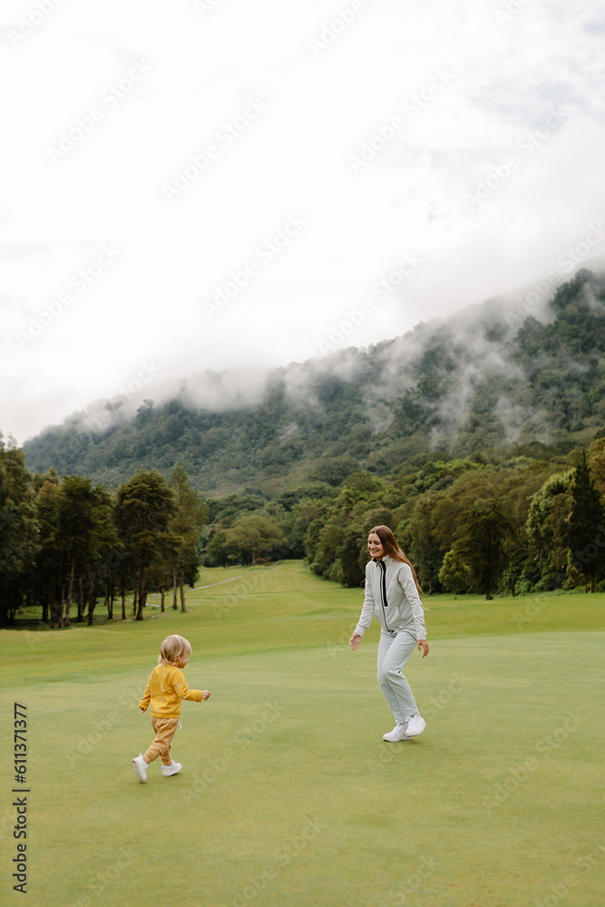 Obraz premium Young woman entertains a child in nature on a green field with a beautiful view of the forest and mountains. Family outdoor recreation happy family