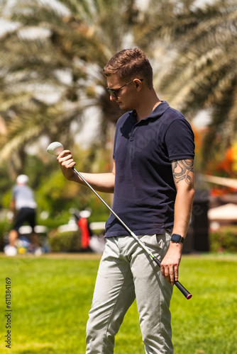 Young guy golfer holding golf club on course in sportswear and sunglasses, training in golf club at palm trees background. Healthy sports lifestyle and leisure activity concept. Copy ad text space