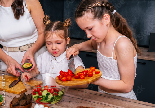 A little girl is preparing a vegetarian salad and adding chopped tomatoes to a bowl together with her younger sister under the guidance of her mothe