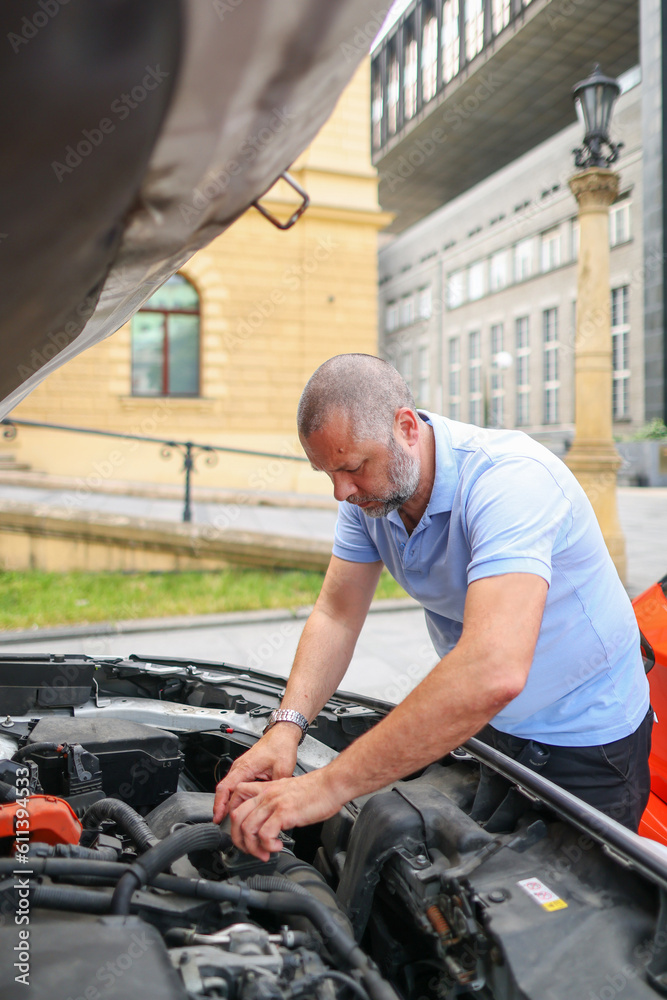 A bearded man leaned over the open hood of a black car with a screwdriver during the repair process. A bearded man leaned over the open hood of a black car with a screwdriver during the repair process.