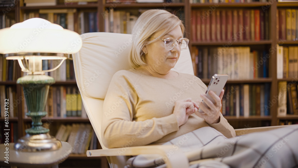 Woman scrolling through web pages on her smartphone, sitting in a cozy armchair in her home office Woman scrolling through web pages on her smartphone, sitting in a cozy armchair in her home office