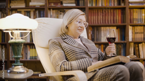 Senior woman reading a book and drinking wine, relaxing in her home library