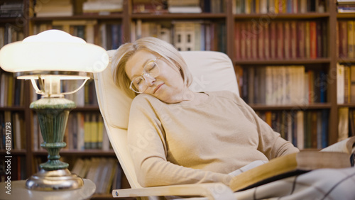 Senior woman falling asleep while reading a book in her library, late evening