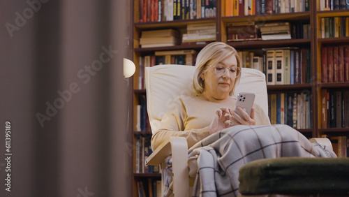 Smiling woman in her 50s covered in blanket scrolling smartphone at home library