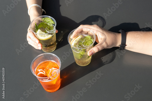 Topview of female hands holding drinks on a grey table; Spritz and hugo in plastic glass; sunlight and shadows in the summertime