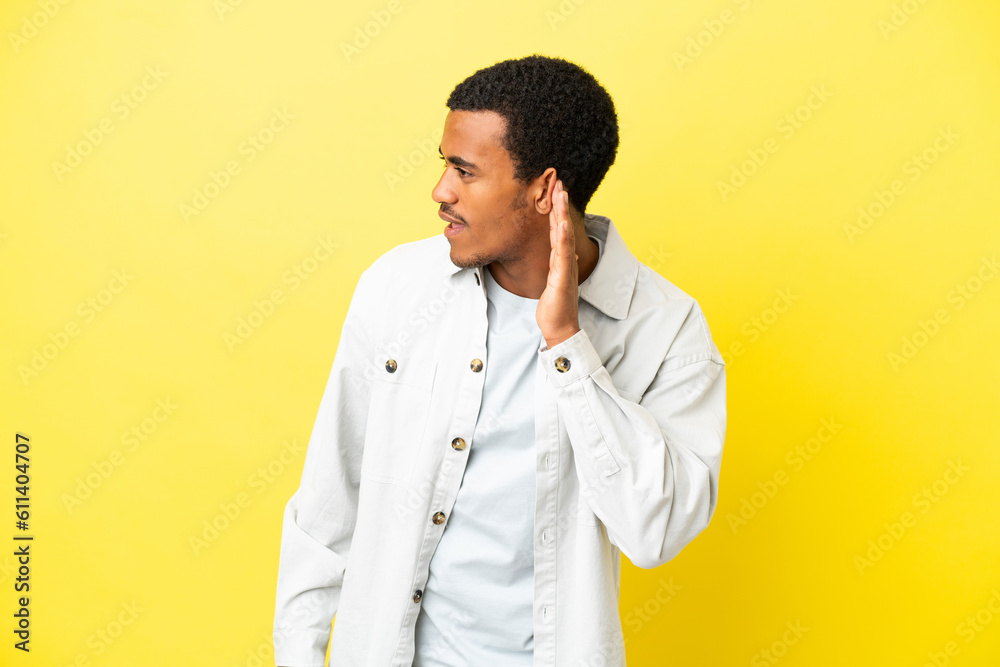 African American handsome man on isolated yellow background listening to something by putting hand on the ear
