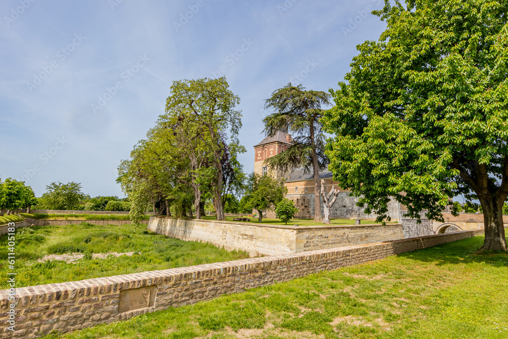 Outer garden of Borgharen castle against blue sky, surrounded by a dry moat, yellowish green grass, trees and building in background, sunny spring day in South Limburg, Netherlands