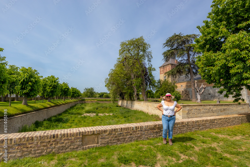 Garden of Borgharen castle with a woman tourist posing happily, surrounded by a dry moat, yellowish green grass, trees and buildings in background, sunny spring day in South Limburg, the Netherlands