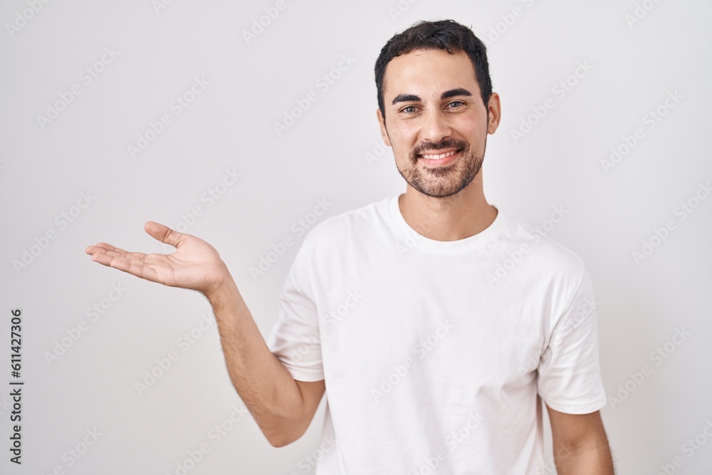 Handsome hispanic man standing over white background smiling cheerful presenting and pointing with palm of hand looking at the camera. Handsome hispanic man standing over white background smiling cheerful presenting and pointing with palm of hand looking at the camera.