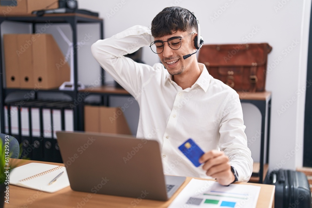 Young hispanic man working using computer laptop holding credit card smiling confident touching hair with hand up gesture, posing attractive and fashionable Young hispanic man working using computer laptop holding credit card smiling confident touching hair with hand up gesture, posing attractive and fashionable