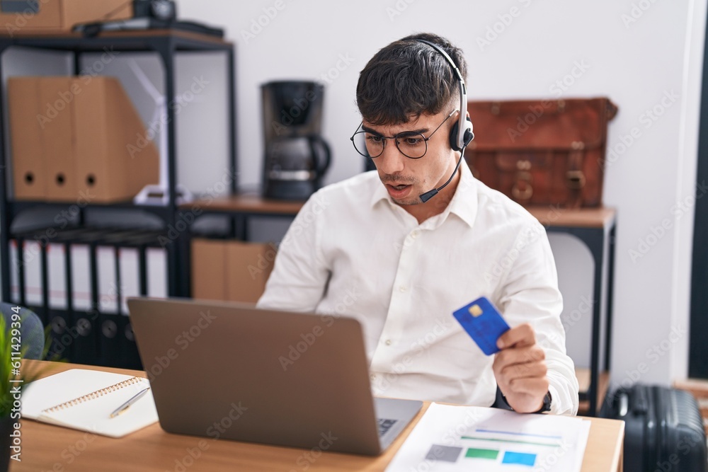Young hispanic man working using computer laptop holding credit card in shock face, looking skeptical and sarcastic, surprised with open mouth Young hispanic man working using computer laptop holding credit card in shock face, looking skeptical and sarcastic, surprised with open mouth