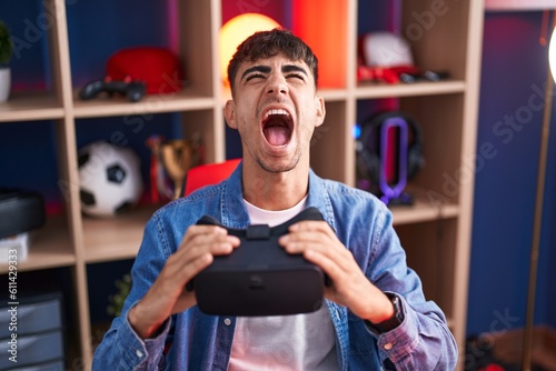 Young hispanic man wearing virtual reality glasses angry and mad screaming frustrated and furious, shouting with anger looking up. photo