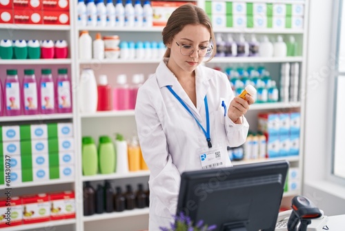 Young blonde woman pharmacist using computer holding pills bottle at pharmacy