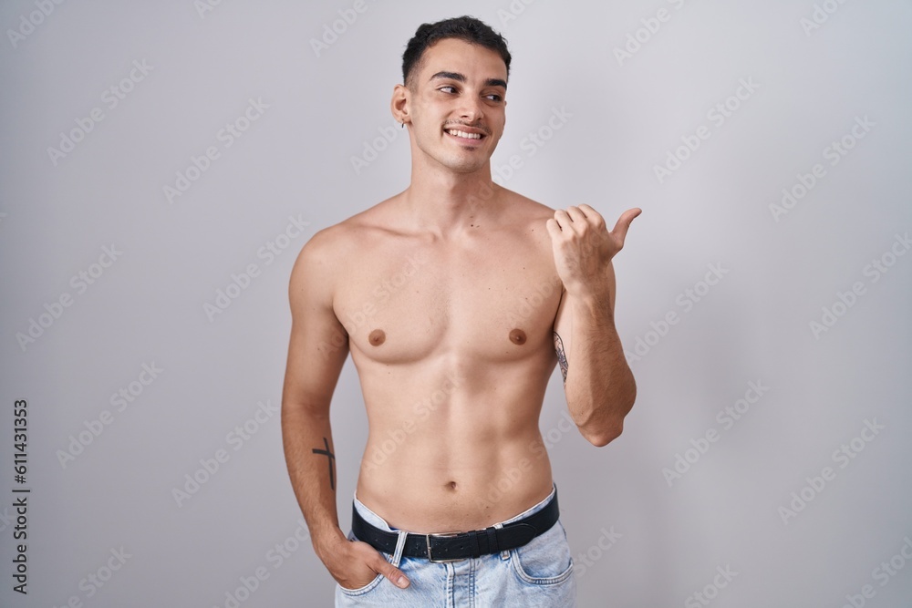 Handsome hispanic man standing shirtless smiling with happy face looking and pointing to the side with thumb up. Handsome hispanic man standing shirtless smiling with happy face looking and pointing to the side with thumb up.