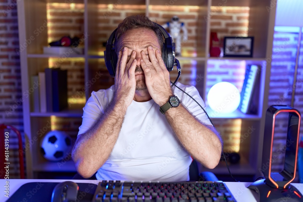 Middle age man with beard playing video games wearing headphones rubbing eyes for fatigue and headache, sleepy and tired expression. vision problem Middle age man with beard playing video games wearing headphones rubbing eyes for fatigue and headache, sleepy and tired expression. vision problem