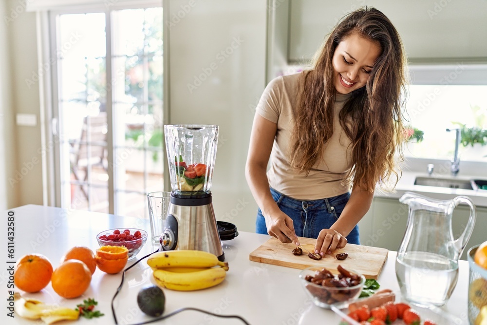 Young beautiful hispanic woman preparing vegetable smoothie with blender cutting date at the kitchen