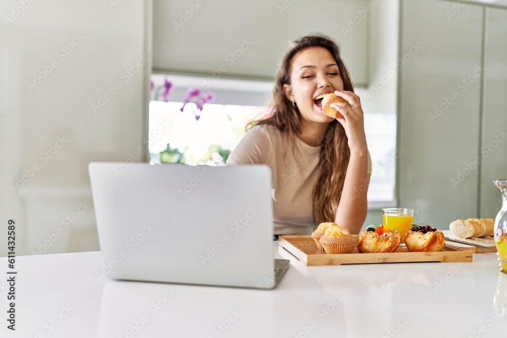 Young beautiful hispanic woman having breakfast using laptop at the kitchen