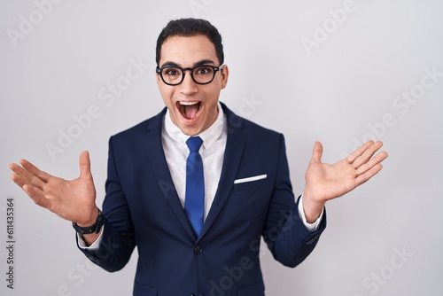 Young hispanic man wearing suit and tie celebrating crazy and amazed for success with arms raised and open eyes screaming excited. winner concept