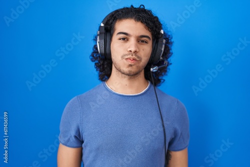 Hispanic man with curly hair listening to music using headphones puffing cheeks with funny face. mouth inflated with air, crazy expression.
