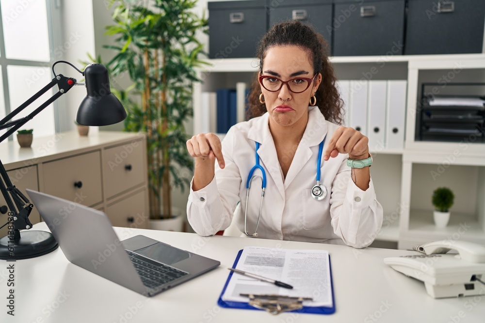 Young hispanic woman wearing doctor uniform and stethoscope pointing down looking sad and upset, indicating direction with fingers, unhappy and depressed. Young hispanic woman wearing doctor uniform and stethoscope pointing down looking sad and upset, indicating direction with fingers, unhappy and depressed.