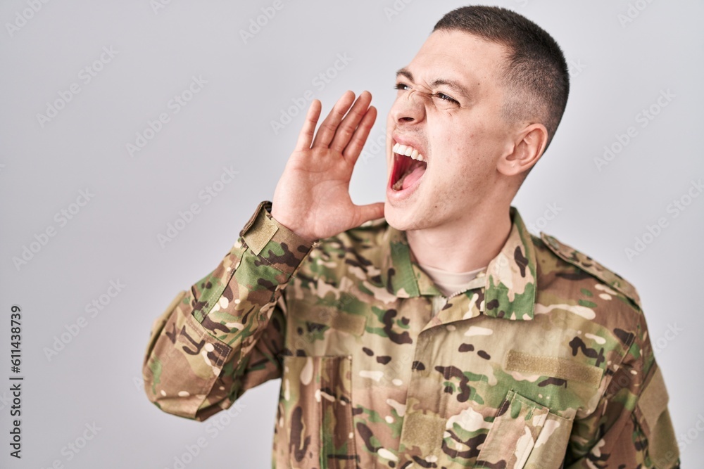 Young man wearing camouflage army uniform shouting and screaming loud to side with hand on mouth. communication concept. Young man wearing camouflage army uniform shouting and screaming loud to side with hand on mouth. communication concept.
