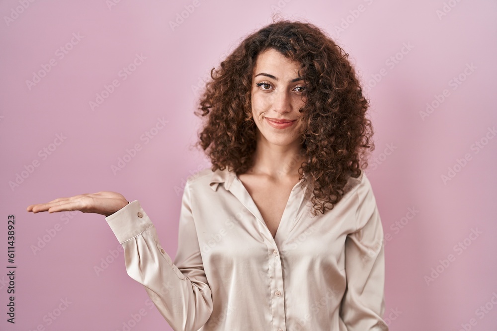 Hispanic woman with curly hair standing over pink background smiling cheerful presenting and pointing with palm of hand looking at the camera. Hispanic woman with curly hair standing over pink background smiling cheerful presenting and pointing with palm of hand looking at the camera.