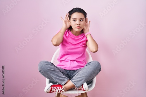Hispanic young woman sitting on chair over pink background trying to hear both hands on ear gesture, curious for gossip. hearing problem, deaf © Krakenimages.com