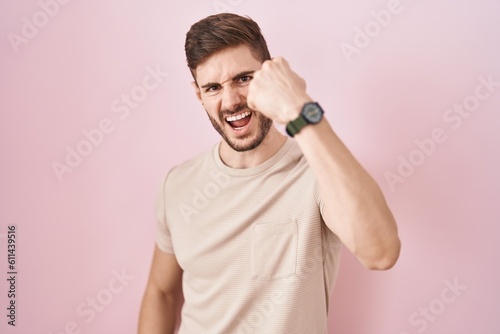 Hispanic man with beard standing over pink background angry and mad raising fist frustrated and furious while shouting with anger. rage and aggressive concept.