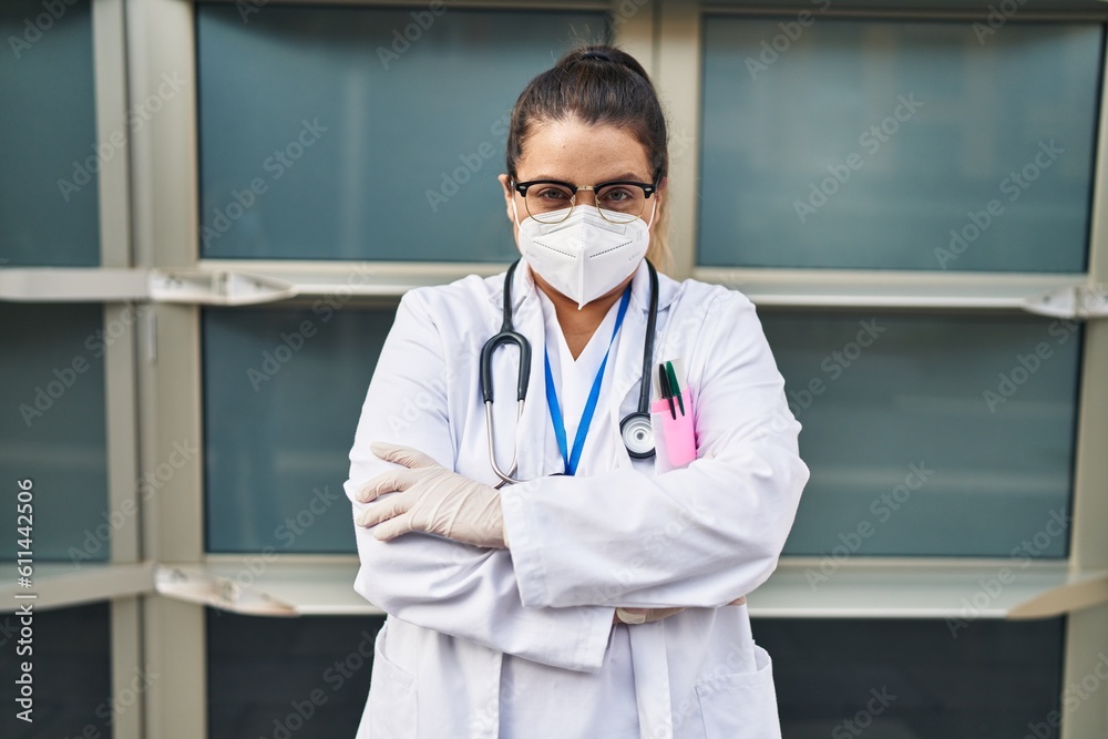 Young beautiful plus size woman doctor wearing medical mask standing with arms crossed gesture at hospital Young beautiful plus size woman doctor wearing medical mask standing with arms crossed gesture at hospital