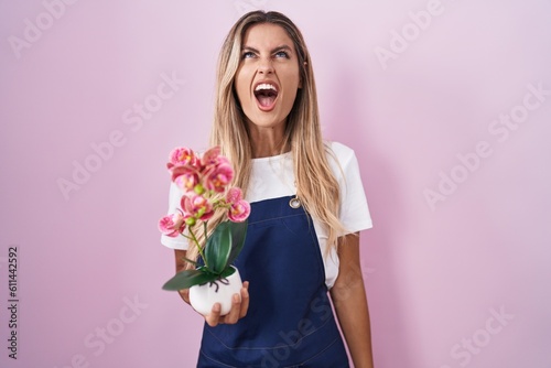 Young blonde woman wearing gardener apron holding plant angry and mad screaming frustrated and furious, shouting with anger. rage and aggressive concept.