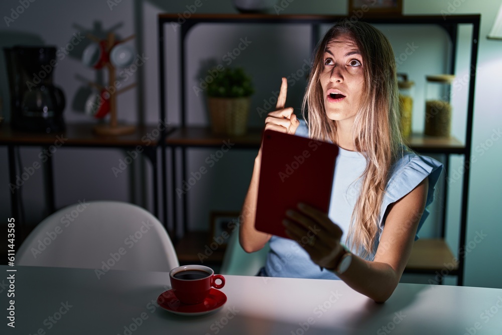 Young hispanic woman using touchpad sitting on the table at night amazed and surprised looking up and pointing with fingers and raised arms. Young hispanic woman using touchpad sitting on the table at night amazed and surprised looking up and pointing with fingers and raised arms.