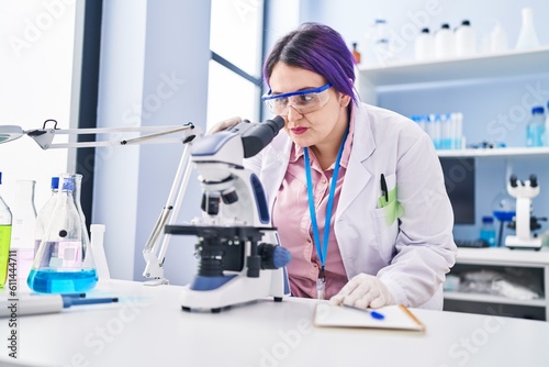 Young beautiful plus size woman scientist writing on notebook using microscope at laboratory