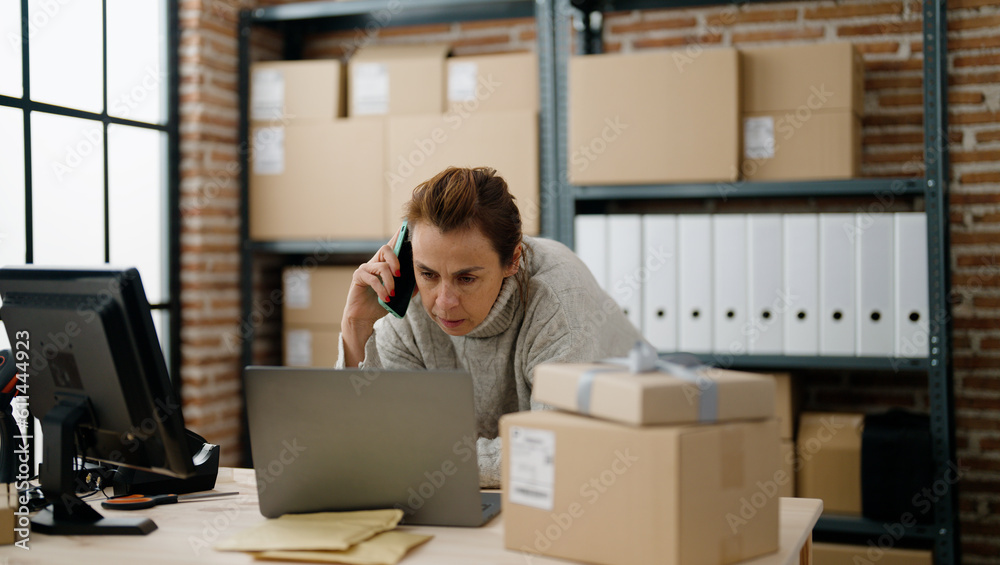 Middle age hispanic woman ecommerce business worker talking on the smartphone using laptop at storehouse office Middle age hispanic woman ecommerce business worker talking on the smartphone using laptop at storehouse office
