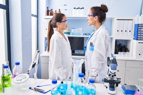 Young mother and daughter at scientist laboratory looking to side  relax profile pose with natural face with confident smile.