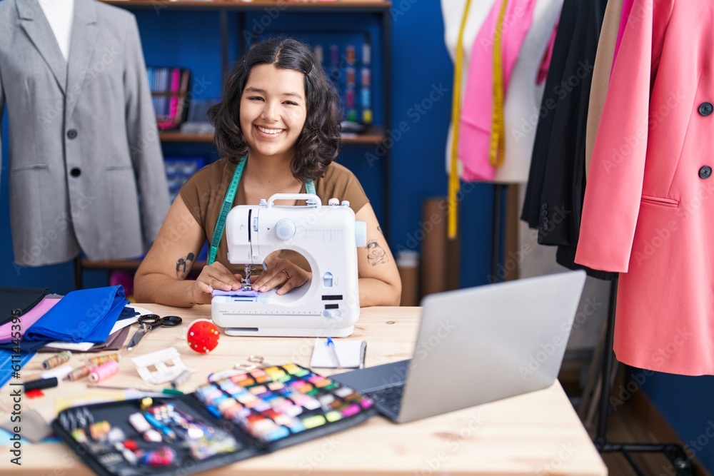 Young woman tailor using sewing machine and laptop at sewing studio