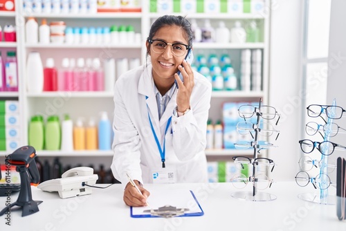 Young beautiful hispanic woman pharmacist talking on smartphone writing on document at pharmacy