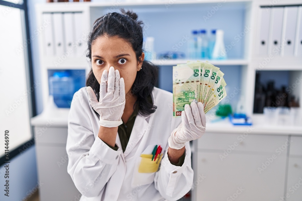 Hispanic woman with dark hair working at scientist laboratory holding money covering mouth with hand, shocked and afraid for mistake. surprised expression Hispanic woman with dark hair working at scientist laboratory holding money covering mouth with hand, shocked and afraid for mistake. surprised expression