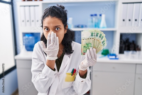 Hispanic woman with dark hair working at scientist laboratory holding money covering mouth with hand  shocked and afraid for mistake. surprised expression